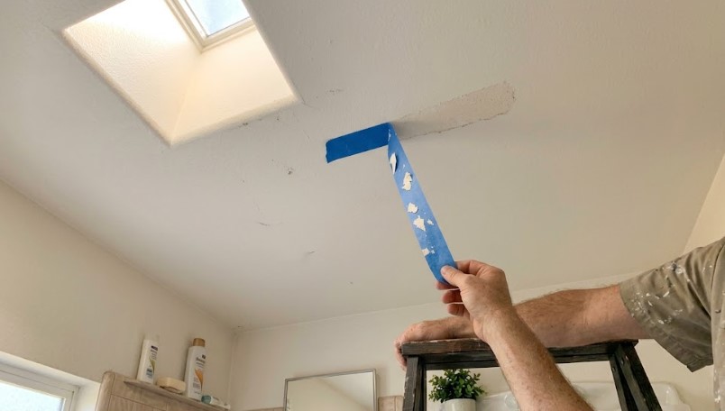 Painter pulling blue tape off a bathroom ceiling with flakes of paint stuck to it, beside a skylight