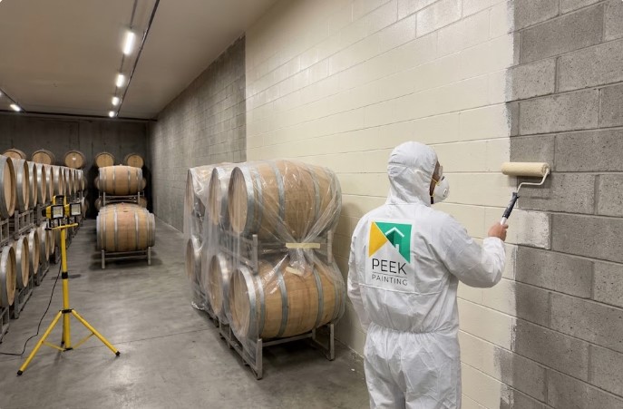 Peek Painting crew member rolling cream coating onto a concrete block wall in a barrel room, next to barrels wrapped in plastic