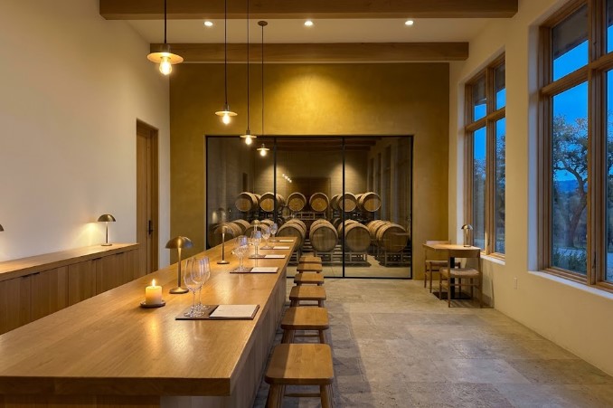 Tasting room in the evening with pendant lights and table lamps glowing over a long oak bar, an ochre wall framing a glass view into the barrel room, and dusk outside the tall windows