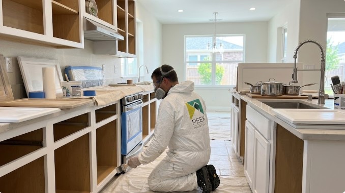 Peek painter painting lower cabinet frames in a masked kitchen