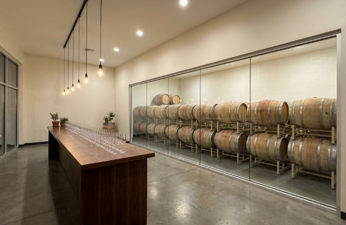 Winery tasting room with a long wooden bar and a glass wall looking into a barrel room with cream-painted block walls