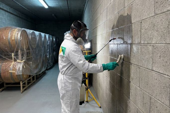 Peek Painting crew member in a protective suit and full-face respirator scrubbing a concrete block wall with a brush and pump sprayer, with barrels wrapped in plastic behind him