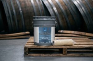 Five-gallon pail of moisture-tolerant coating and a roller cover on a wooden pallet in front of wine barrels