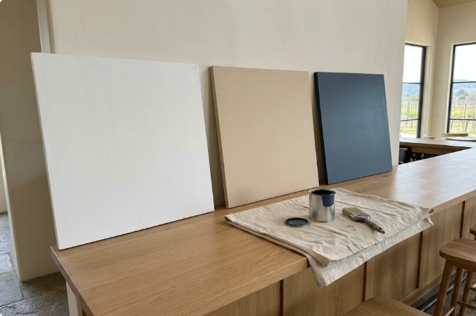 Three large paint sample boards, white, sandy tan and deep navy, leaning against a wall on an oak tasting bar with vineyard views through the windows